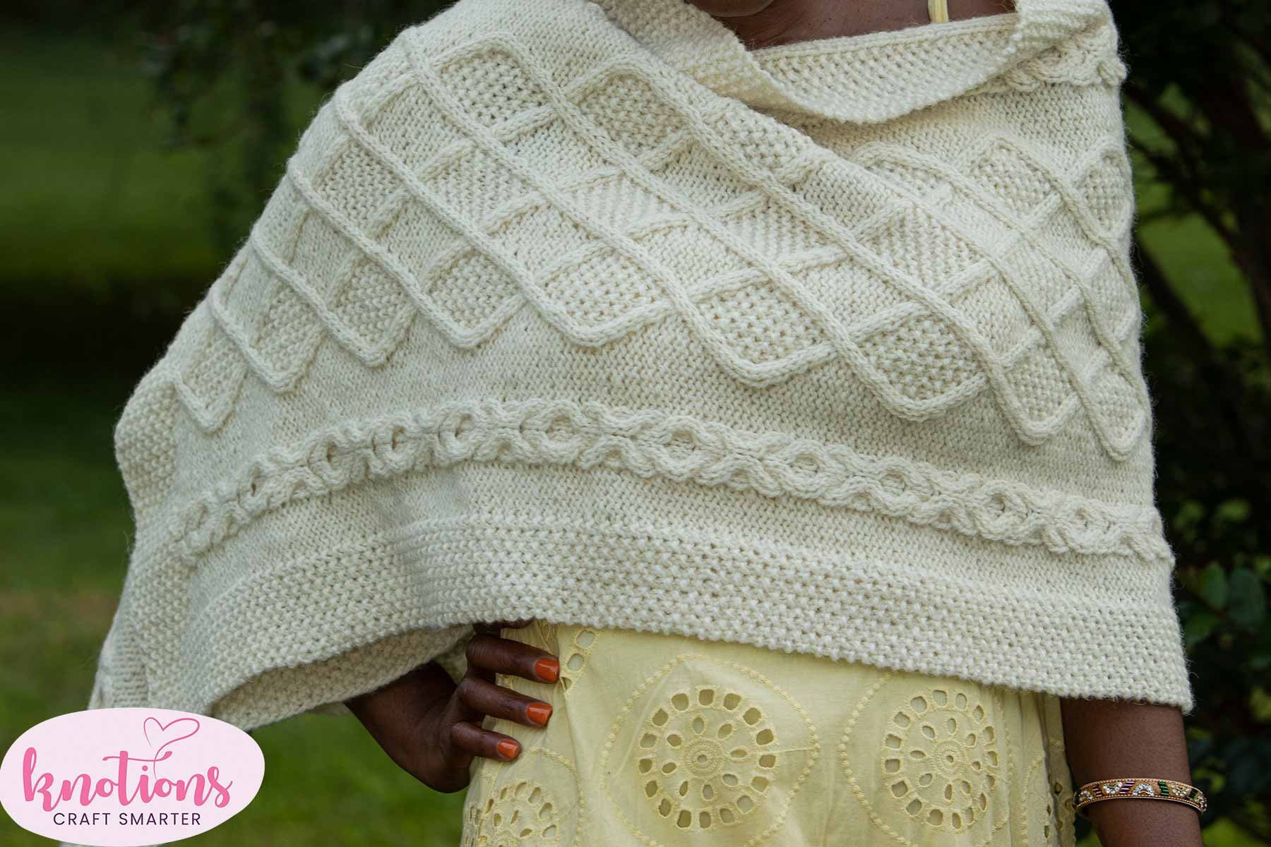 A close up view of the Inishere wrap, a white cabled knitted shawl, modelled by a woman wearing a summer dress.