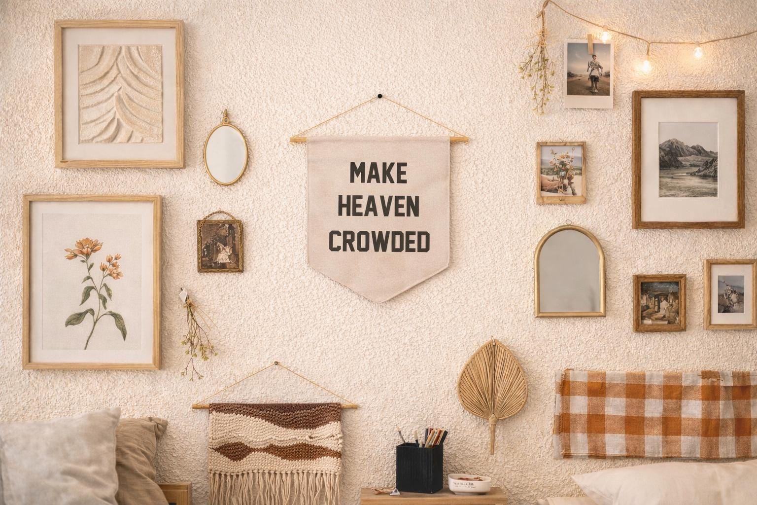 A warm, bohemian-style gallery wall in a bedroom featuring a cream textured wall. At the center hangs a cream felt pennant banner reading MAKE HEAVEN CROWDED in bold black lettering, suspended from a wooden dowel. Surrounding it is an eclectic mix of gold and wood-framed art including a botanical floral print, a leaf texture print, a small vintage portrait, and landscape photographs. Two mirrors — a small oval and a larger arch-shaped mirror — are interspersed among the frames. A strand of warm globe string lights and a sprig of dried flowers hang in the upper right. Below the banner, a woven macramé wall hanging and a dried palm leaf fan add texture. A small nightstand holds a black pen cup and a small dish, with a rust-orange plaid headboard partially visible on the right and neutral pillows on the left.