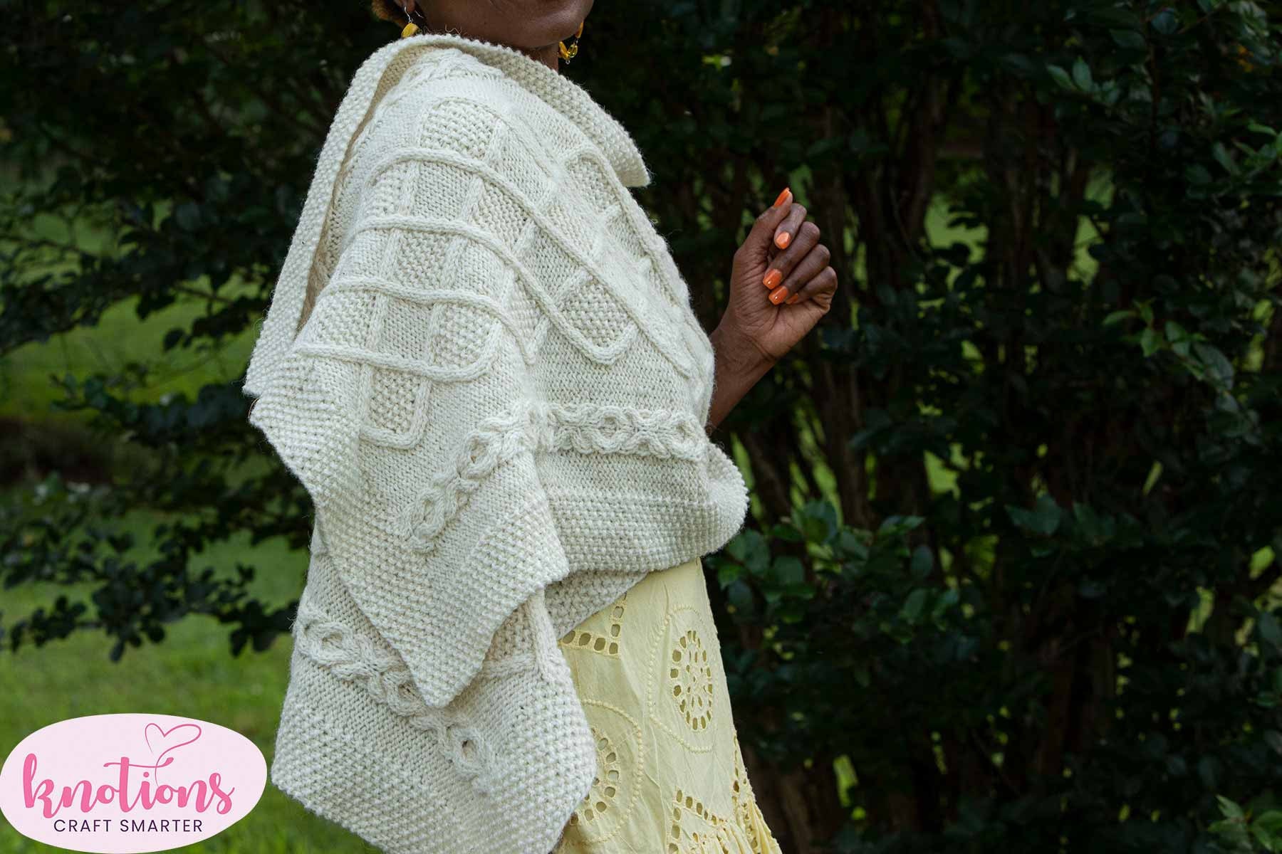 Side view of the Inishere Wrap, a knitted cabled shawl, worn over a summer dress by a smiling woman.