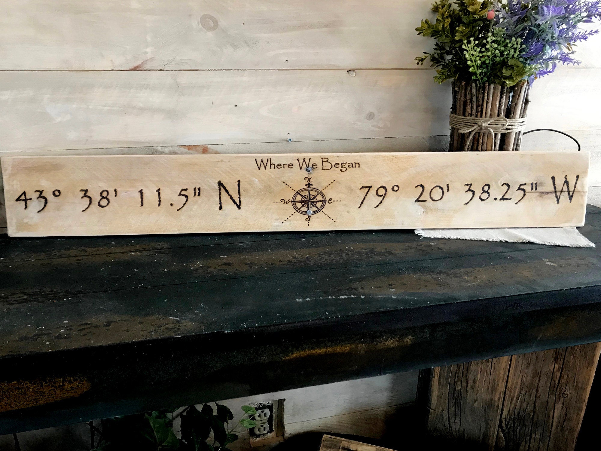 This picture shows a handmade wood location sign. The artwork has been burned into the wood. There is a compass rose in the center and personalized coordinates on either side. The phrase Where we began is burned over the compass.