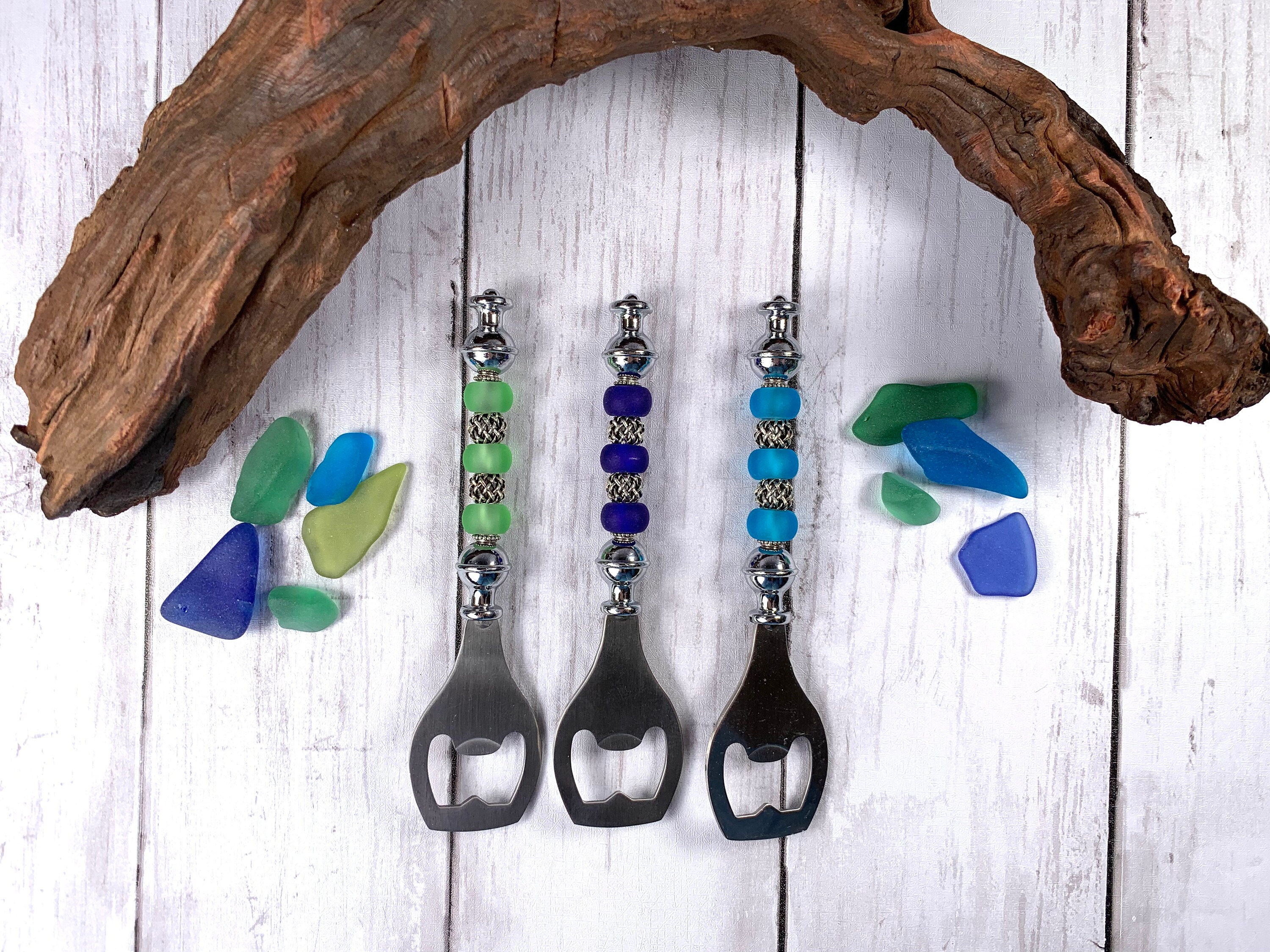 Three beadable bottle openers with colorful beads in shades of green and blue, accented with silver spacer beads. Displayed on a rustic wooden surface with scattered sea glass pieces and a driftwood branch.