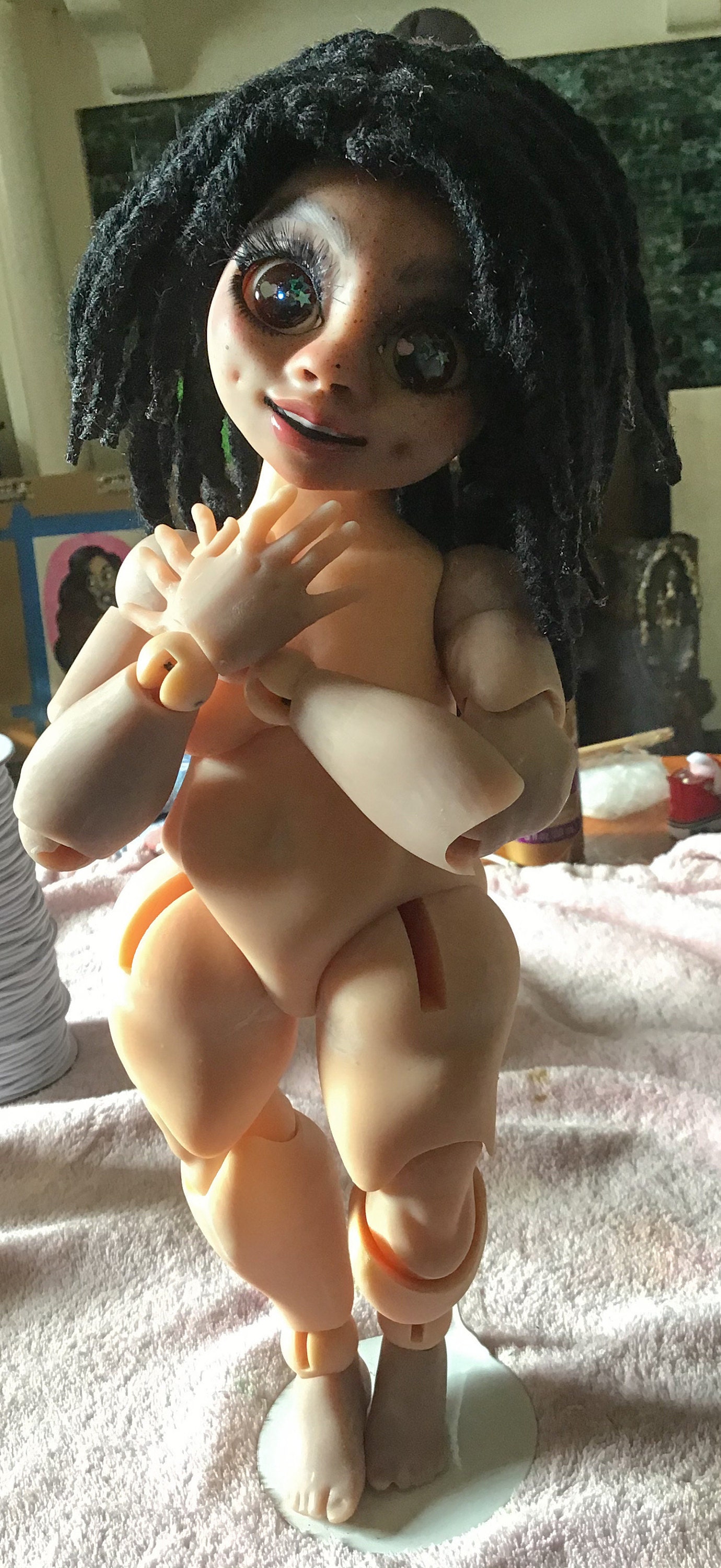 3D printed doll, Nina, is standing. She is positioned with one leg firm on the surface and the other slightly bent at the knee. Both hands cover her chest as they overlap each other.