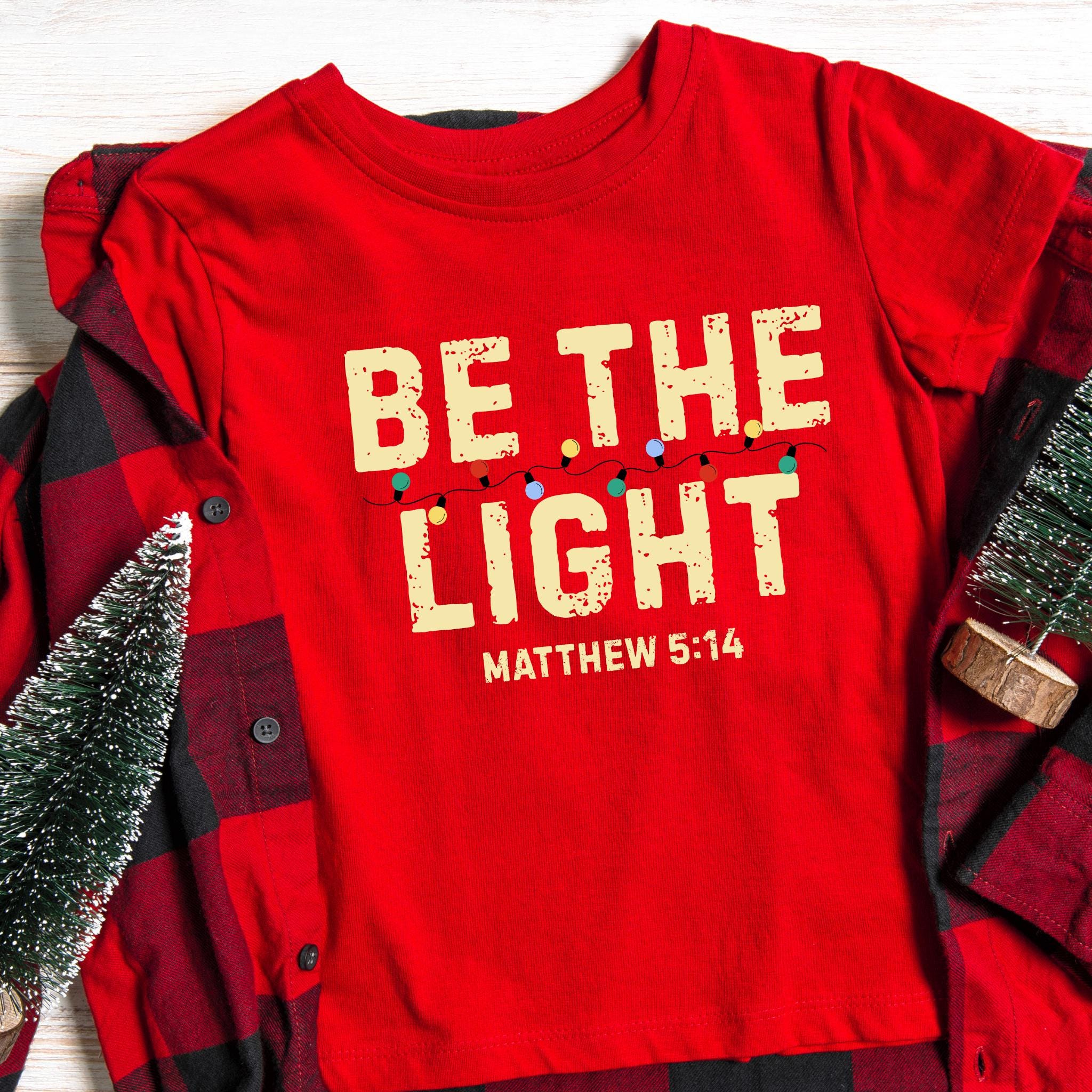 Red Christmas t-shirt with Bible verse Matthew 5:14 ‘Be The Light’ decorated with colorful Christmas lights, Christian holiday faith shirt.