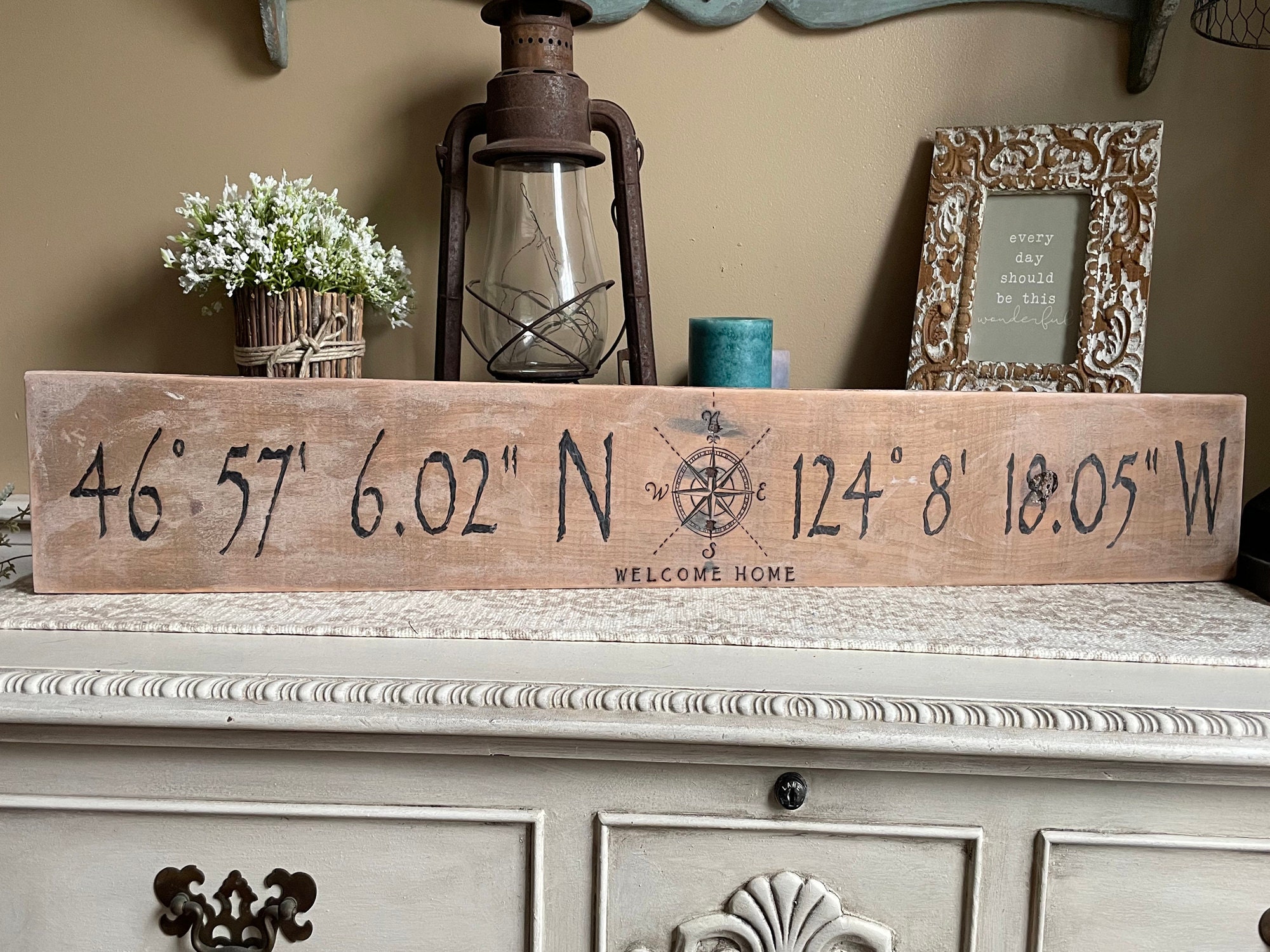 This picture shows a handmade location sign. It is a 36 inches long and 5 inches tall. The personalized coordinates are burned onto the wood. a decorative compass rose is in the center.