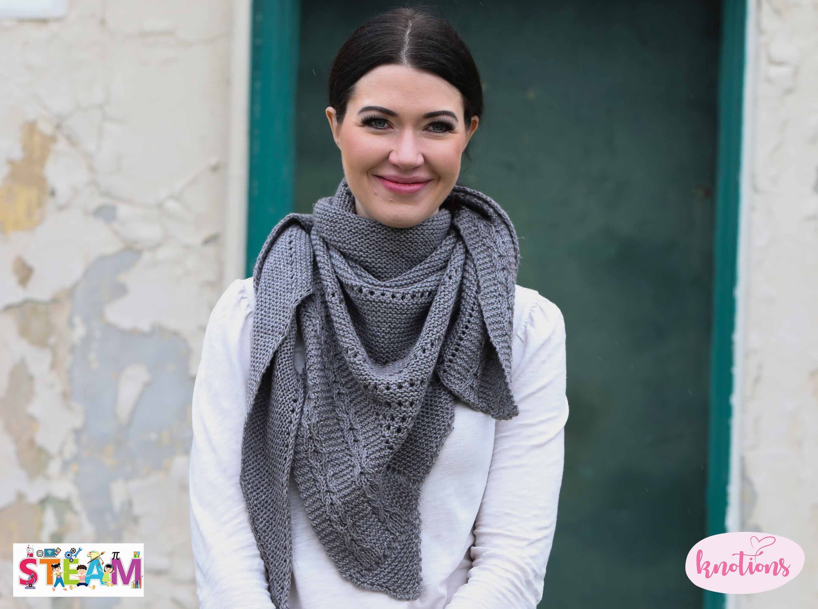 A smiling woman wears a grey knitted shawl, which features a cable pattern along rows of eyelets.