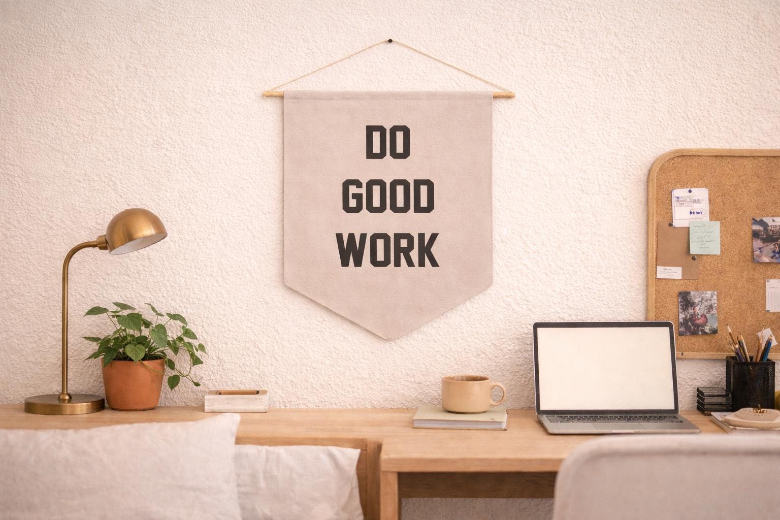 A clean, minimal home workspace with a light textured wall. A cream felt pennant banner reading DO GOOD WORK in bold dark letters hangs from a wooden dowel above the desk. On the light wood desk sits an open laptop with a blank white screen, a ceramic mug resting on a stack of books, a small potted green plant in a terracotta pot, and a brass arc desk lamp. To the right, a cork bulletin board is pinned with notes, cards, and photos, with a black pen and pencil cup and small accessories nearby. A white chair or couch edge is partially visible in the foreground.
