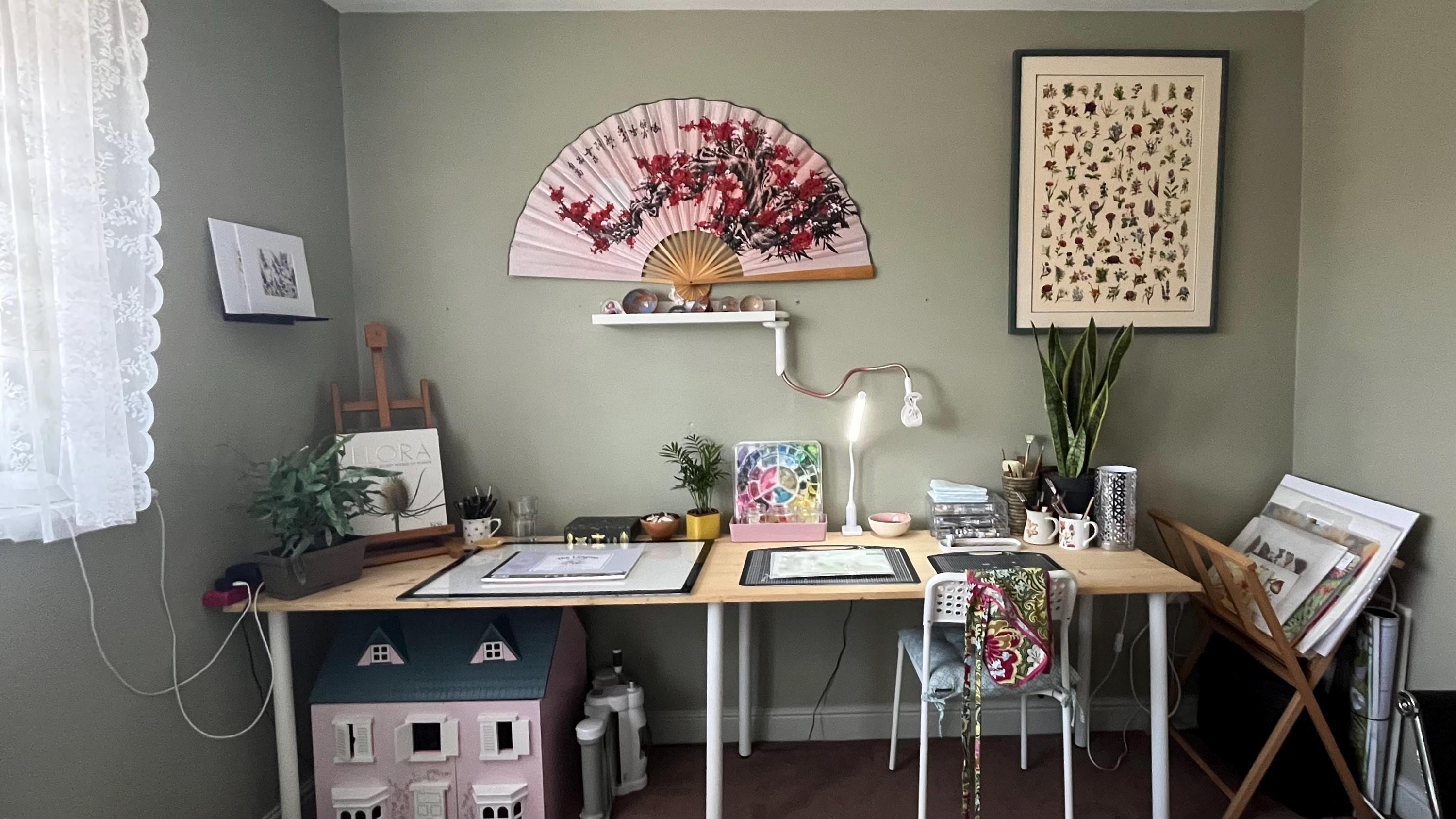 Art Studio with a huge fan on the wall, a framed floral picture and wide desk, chair and a dolls house project under the table end, all by a window for natural light.