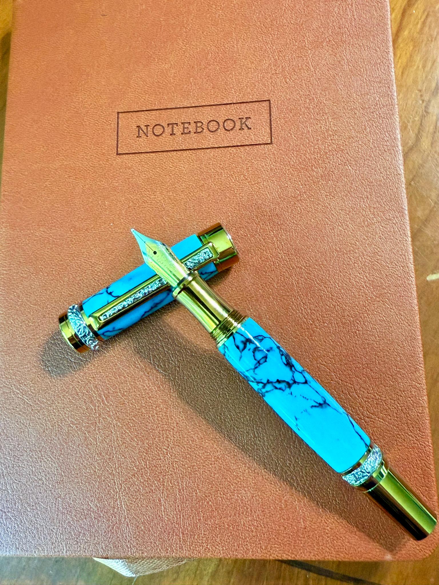 Fountain Pen laying on a leather Journal. The pen has turquoise body with 22K Gold Trim and silver accents. The nib is a medium #6 24k gold with iridium tip