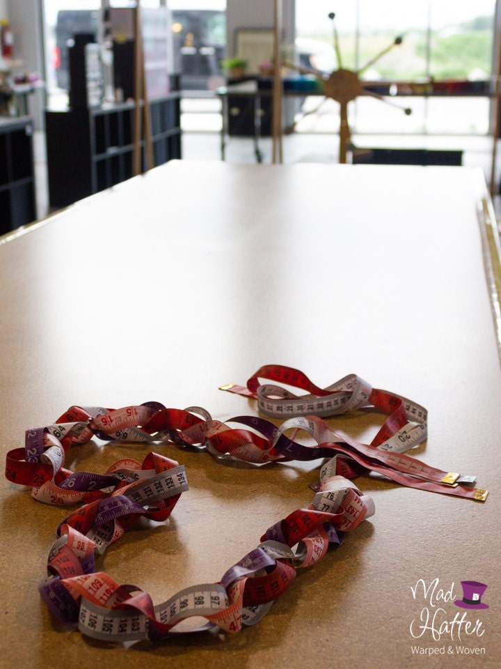 Measuring tapes are entwined together and shaped into a heart laying on a wooden work bench inside Mad hatter Warped & Woven Studio. Blurry in the background, there are large windows with sunshine coming through.