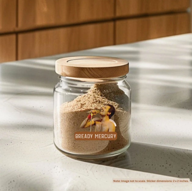 A jar of sourdough starter with a wooden lid, featuring a humorous “Bready Mercury” sticker with an image of Freddie Mercury singing. The jar is sitting on a kitchen counter with natural light streaming over it. The sticker is approximately 2 x 2 inches in size.
