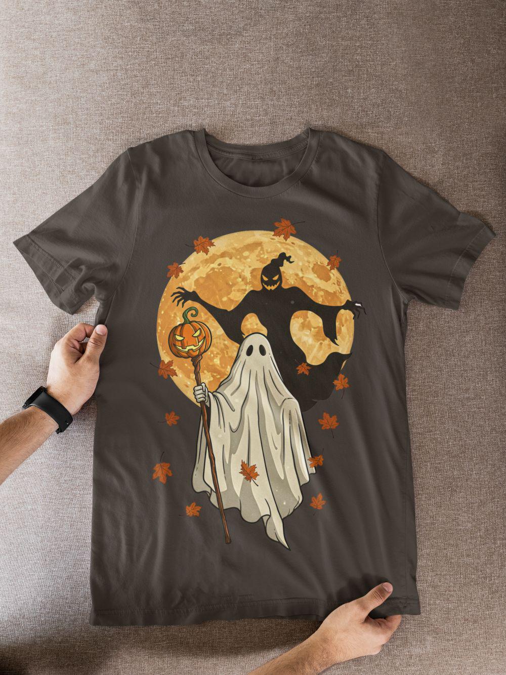 Black unisex T-shirt on a table featuring spooky-cute ghost and full moon design with orange leaves—Halloween fall aesthetic.