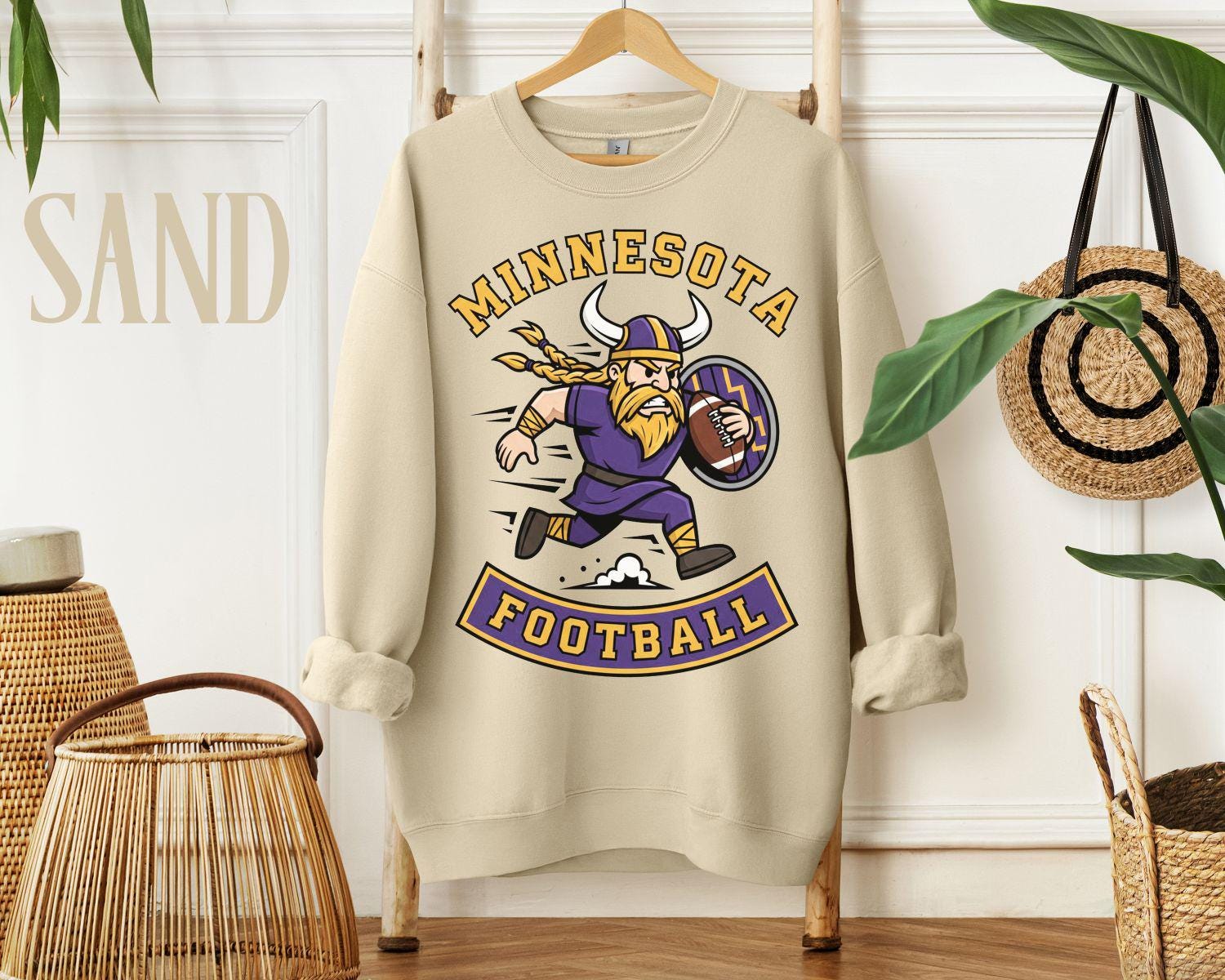 A sand-colored sweatshirt with a retro Minnesota football design featuring a running Viking mascot, styled on a wooden hanger in a bright room.