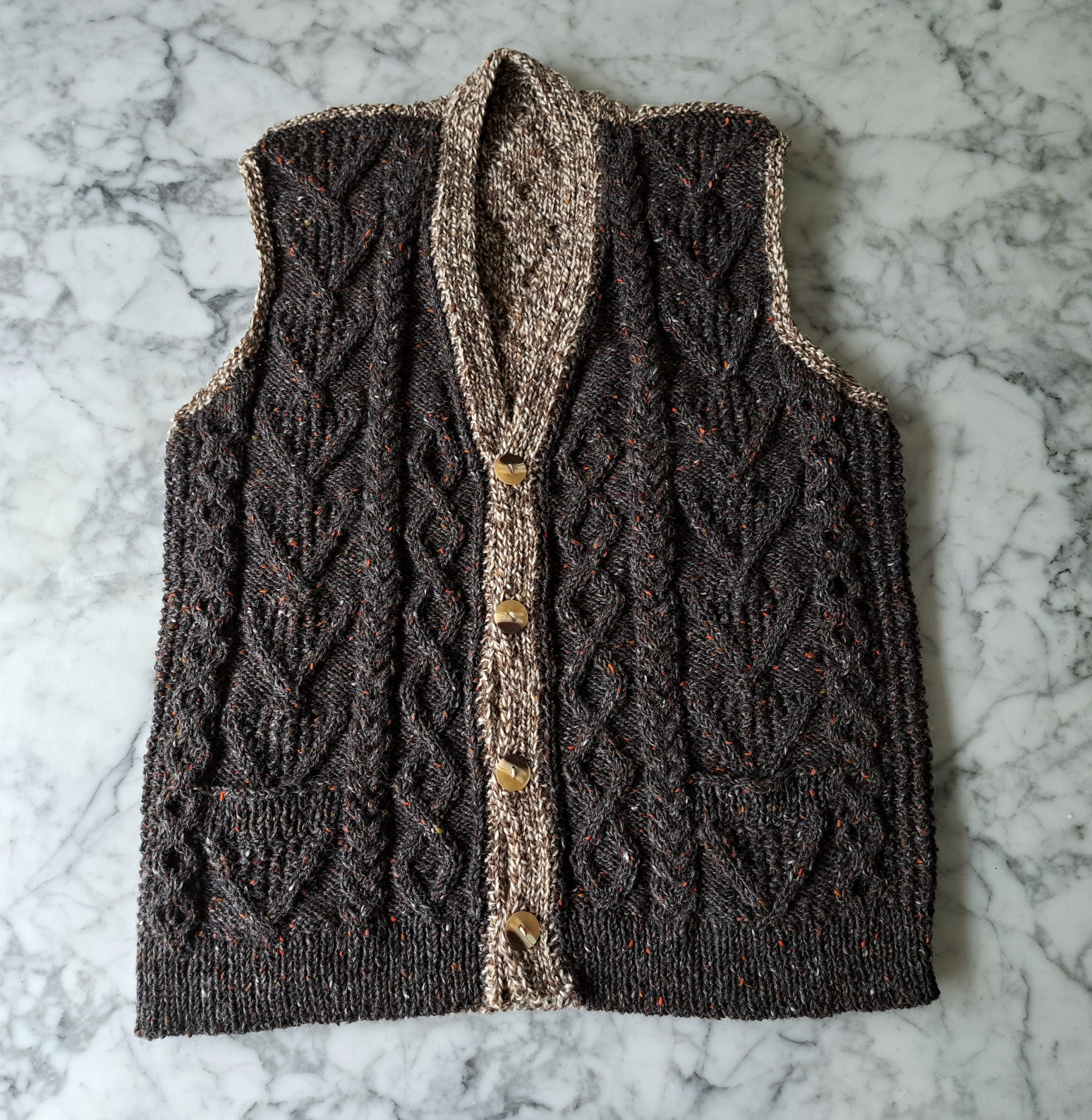 a two-tone Aran knit cabled waistcoat in shades of brown lies flat on a marble background