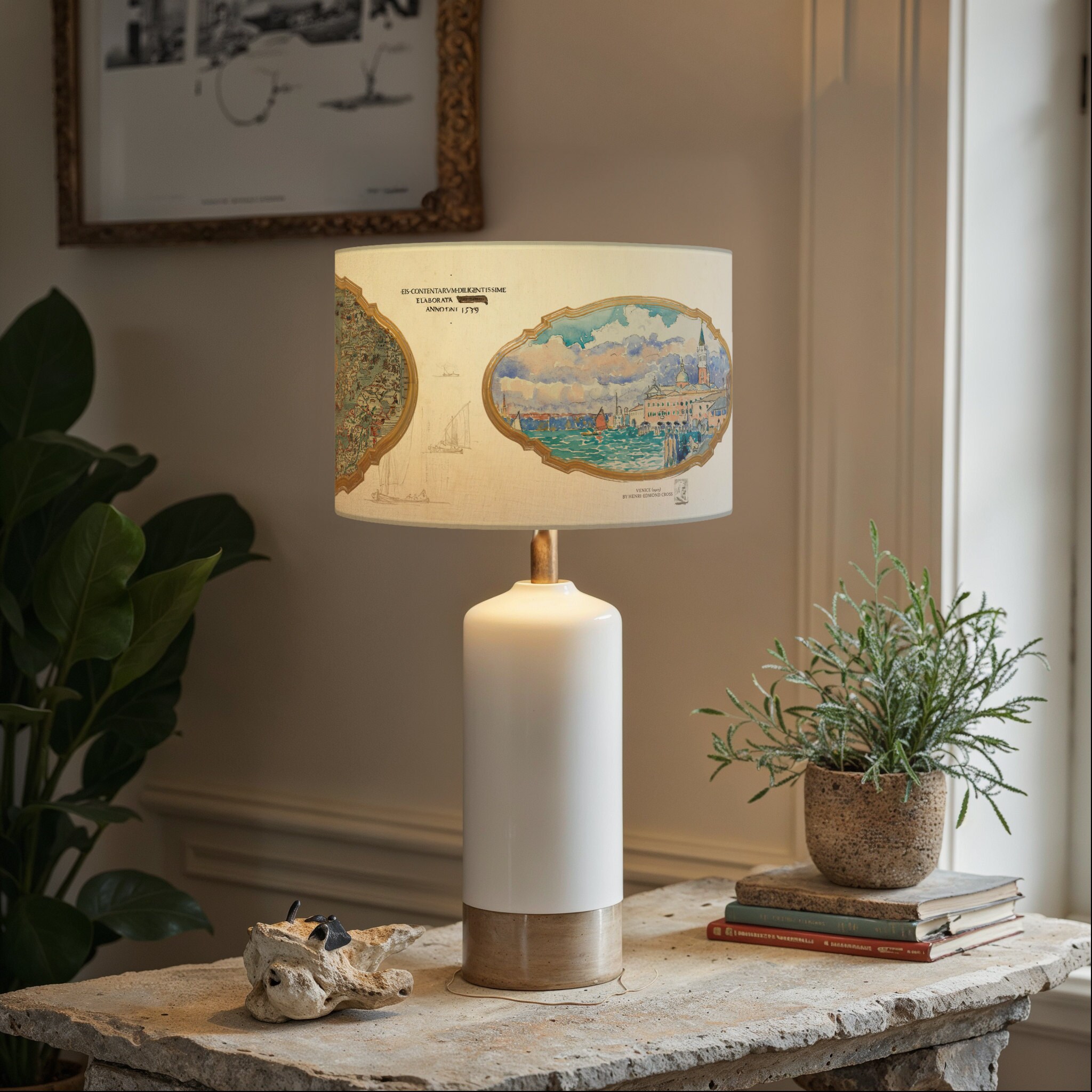 Lampe moderne avec décor de cartes historiques Lampe au pied blanc et doré, avec un abat-jour orné de cartes et dillustrations maritimes vintage. Posée sur une table en pierre brute, entourée de livres anciens et de plantes en pot, la lampe crée un contraste harmonieux entre le style moderne et le charme des illustrations anciennes.