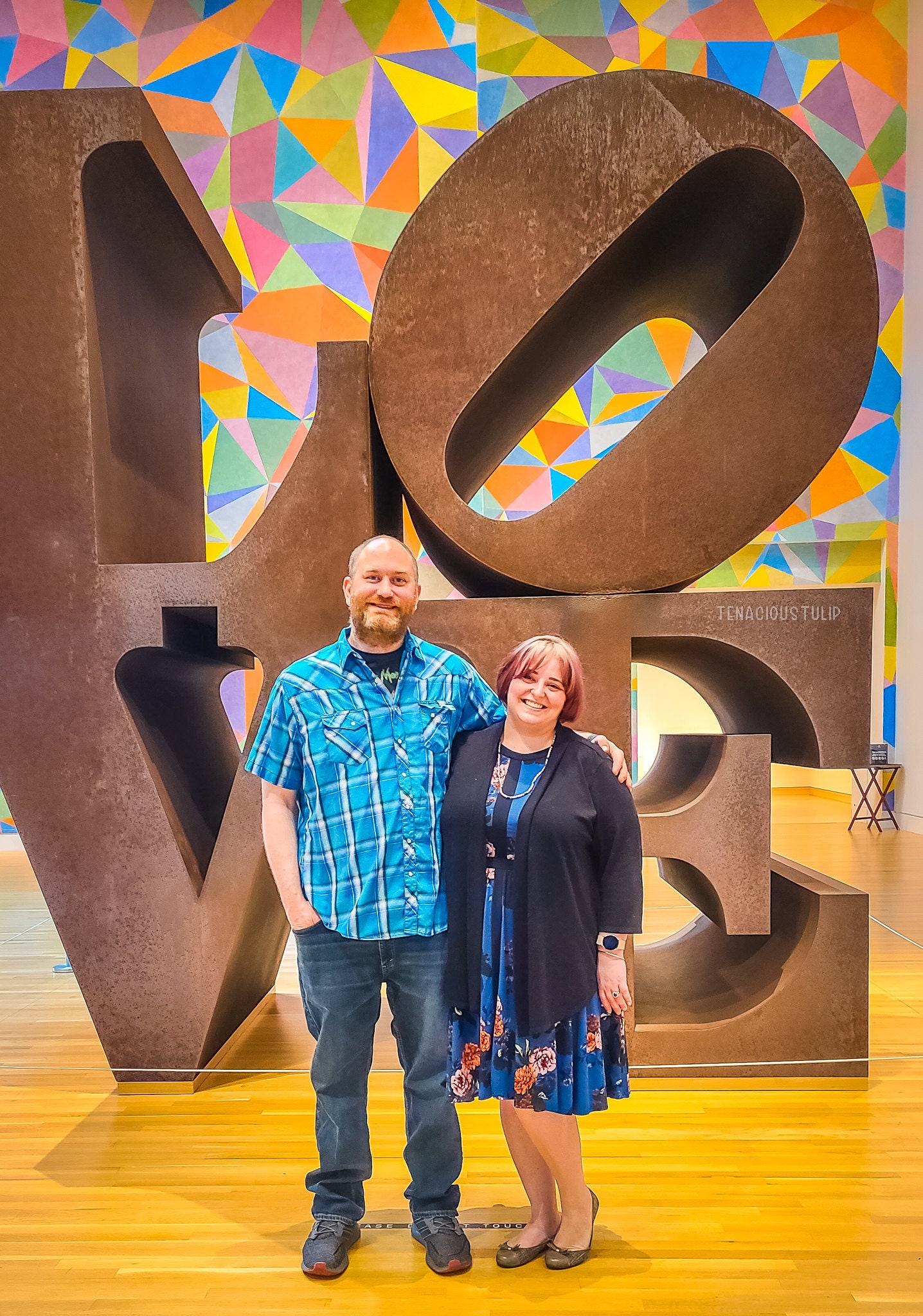 Man in a blue shirt and jeans, woman in a dress and black sweater stand in front of a statue spelling LOVE