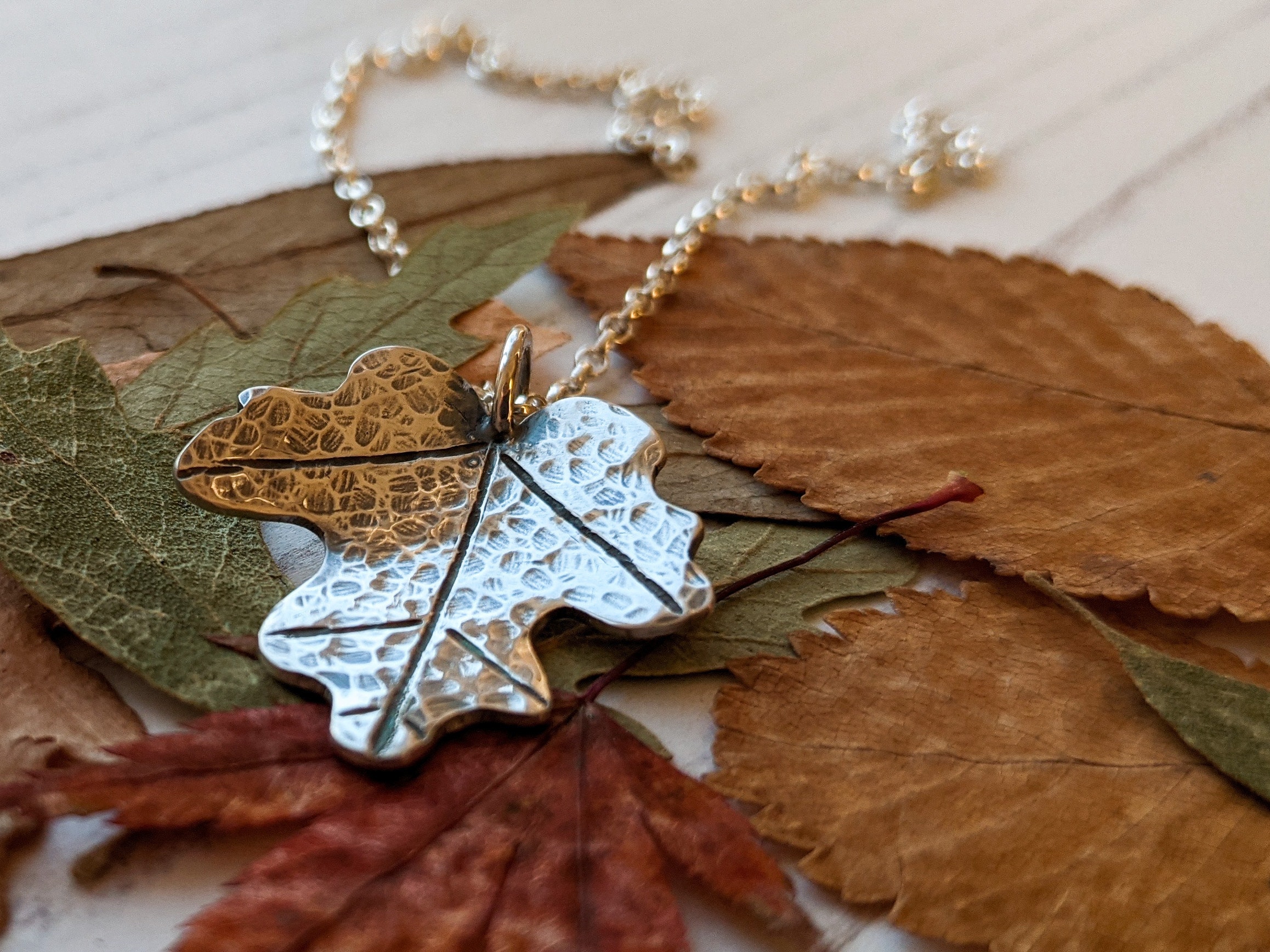 A silver pendant in the form of maple leaves, with a silver chain, against a background of autumn coloured leaves.