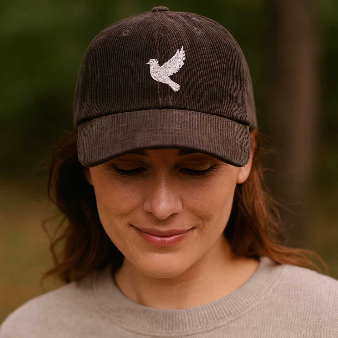 A woman in a light gray sweatshirt wears a dark brown corduroy cap with a white dove embroidered on the front. She looks down with a soft smile in an outdoor forest setting.