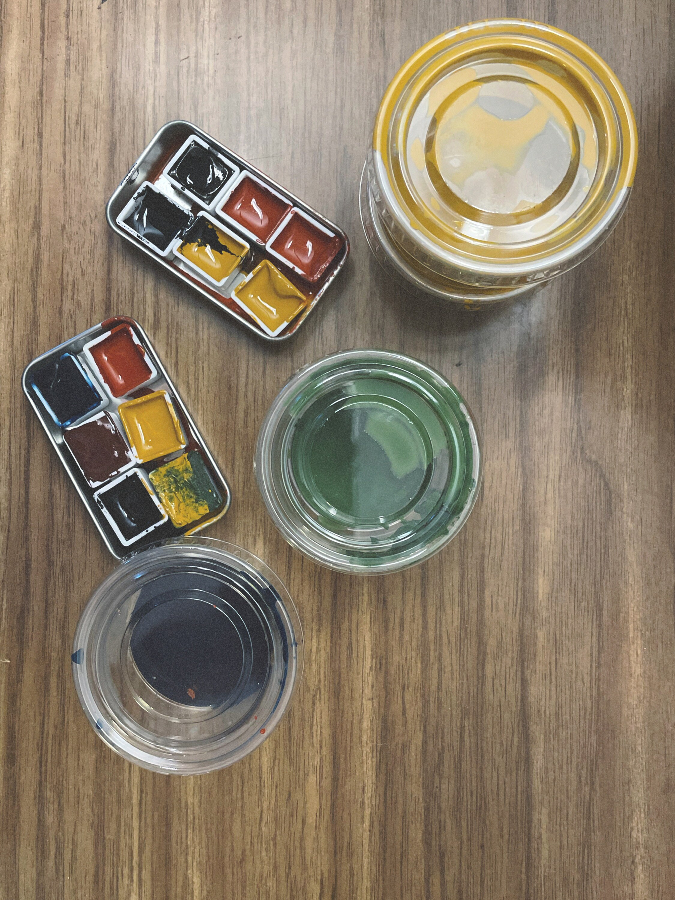 Two small metal containers of finished watercolors, each holding six paint pans. The bottom container has Maya Blue, Red and Yellow Ochre, Armenian Purple Ochre, French Green Clay, and Bone Black. The second container has two each of Red Ochre, Yellow Ochre, and Bone Black. To the right of these containers are small plastic dressing containers with inks - one in Maya Blue, one French Green Clay, and two Yellow Ochre.