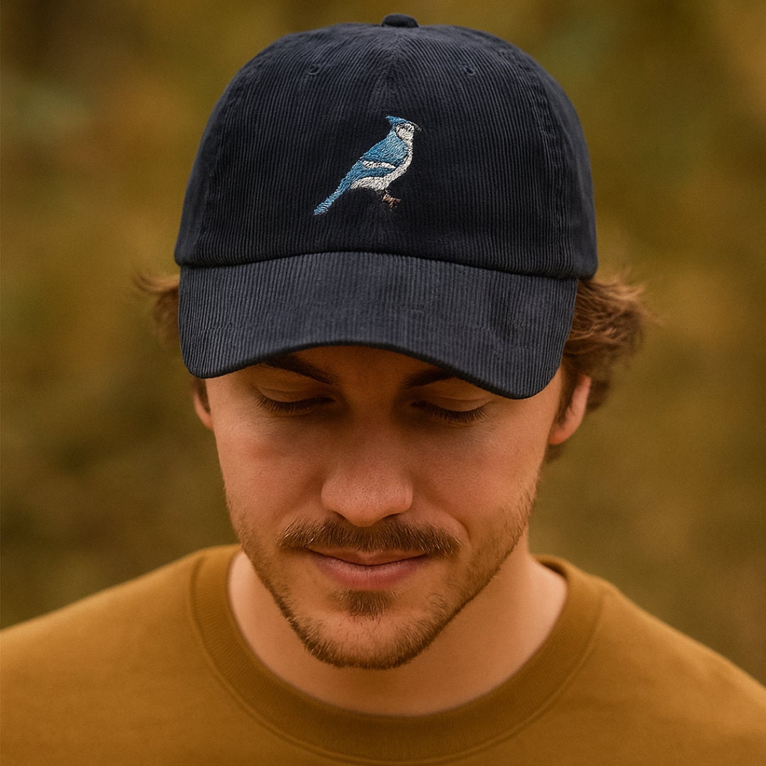 A man wearing a navy blue corduroy baseball cap embroidered with a blue jay, looking down with a gentle smile. The background is softly blurred with autumn tones.