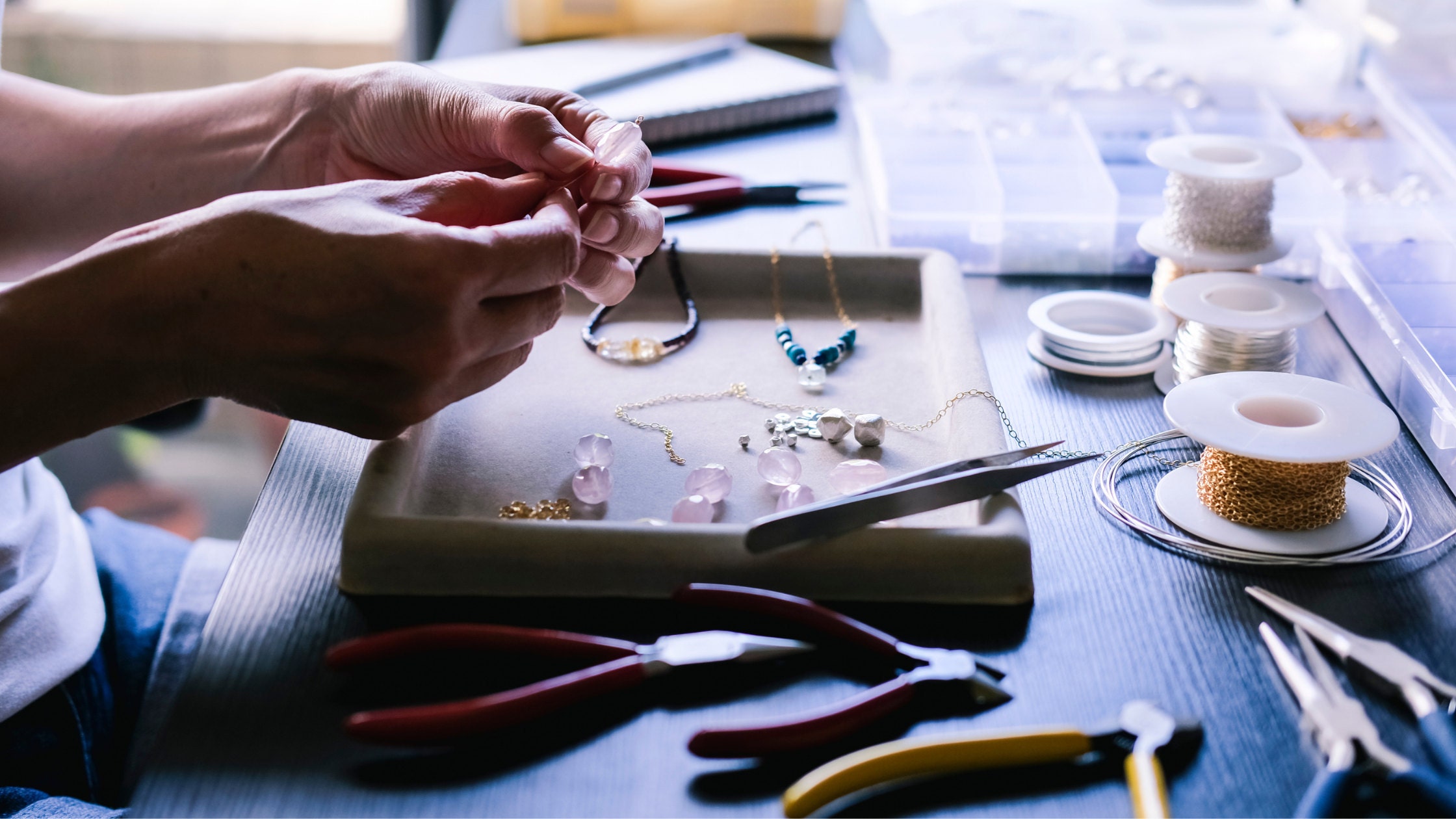 A photo of hands creating jewelry. A jewelers bench surrounded by jewelry making supplies.
