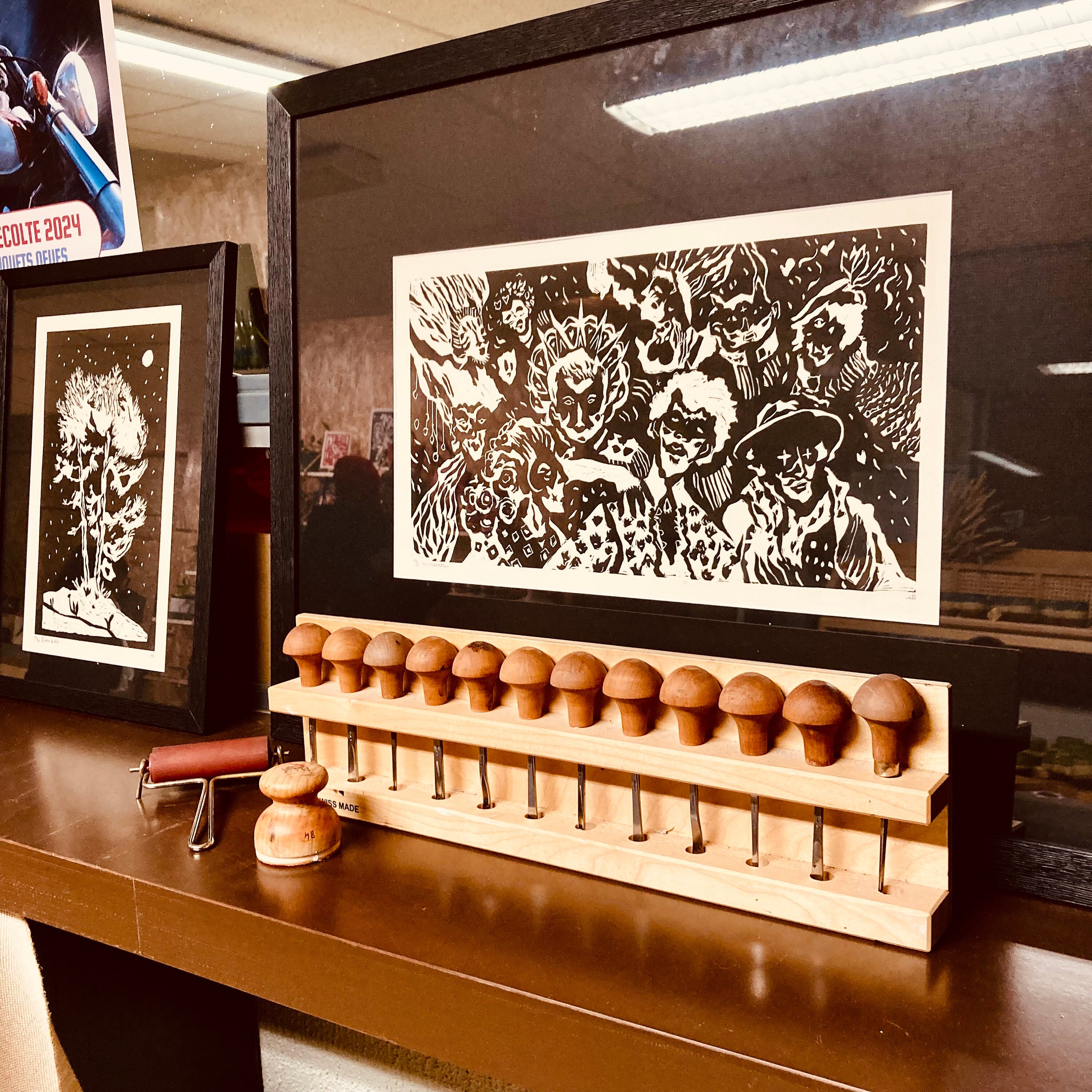 Outils utiles pour réaliser des Linogravures, présentés devant une création encadrée en noir et blanc, représentant plusieurs portraits de personnages masqués.