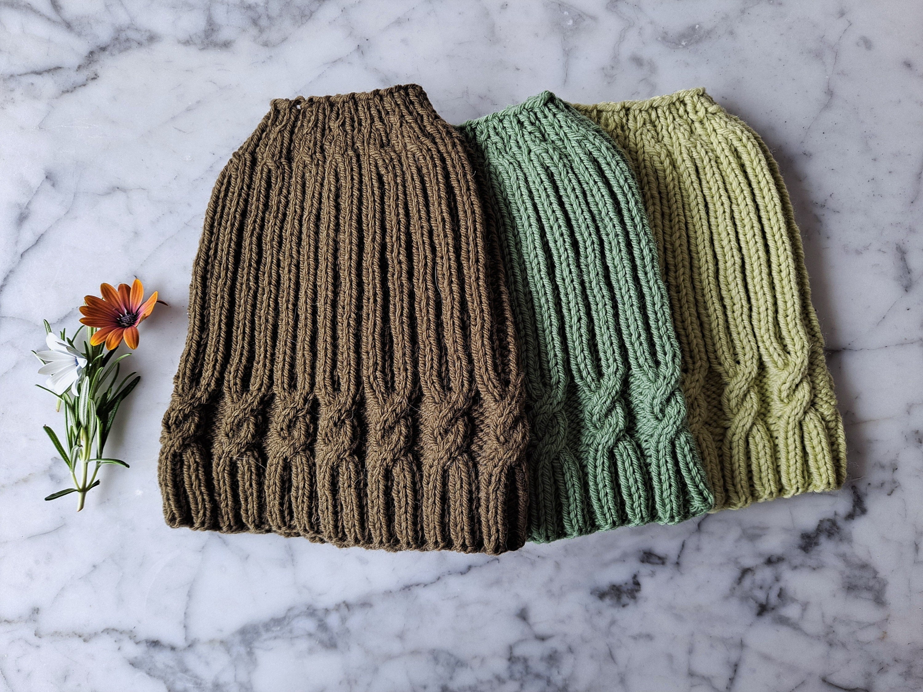 3 hats with a simple cable pattern lie flat on a marble background; the hats are coloured brown and green.