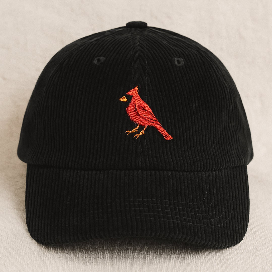 A close-up of a black corduroy baseball cap featuring a detailed red cardinal embroidery, photographed against a soft, neutral background.