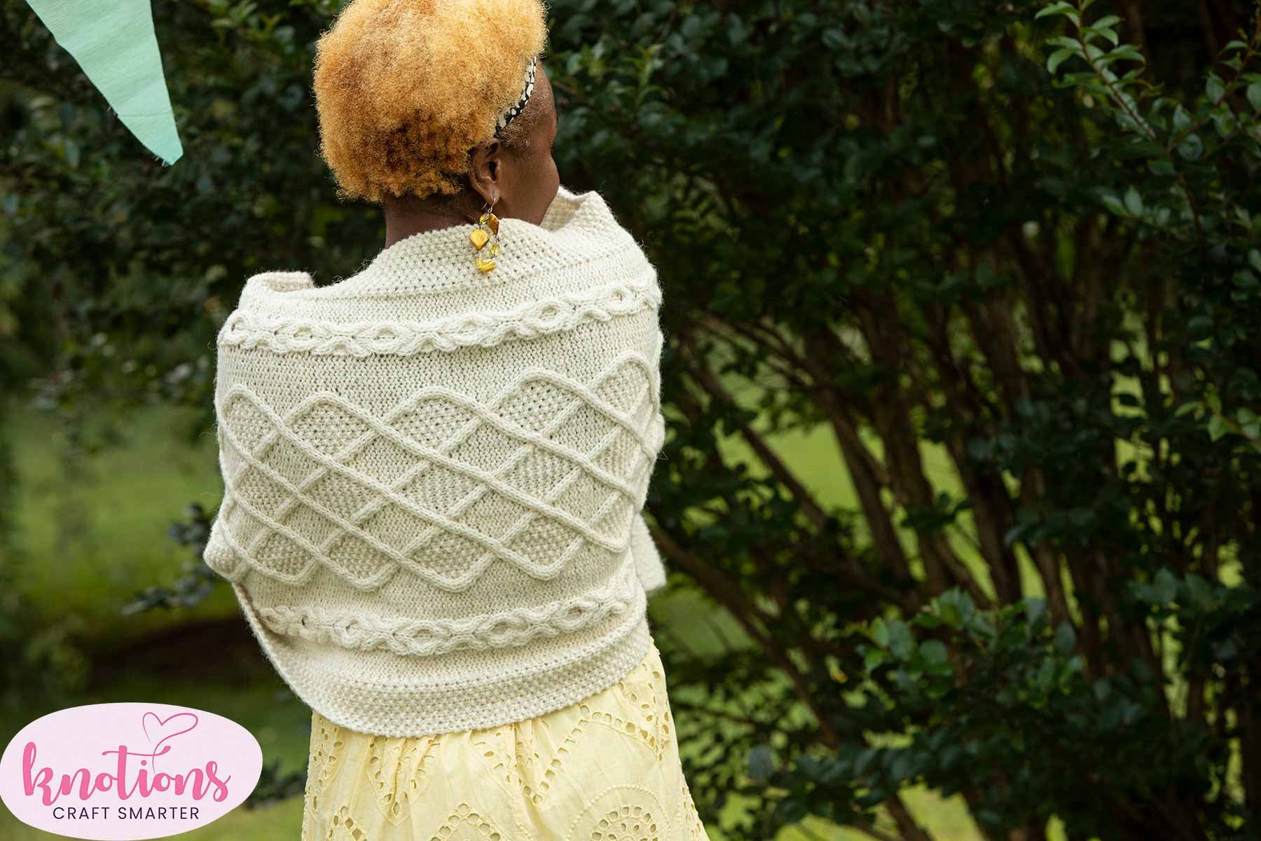A back view of the Inishere Wrap, a white cabled knitted shawl, worn with a summer dress.