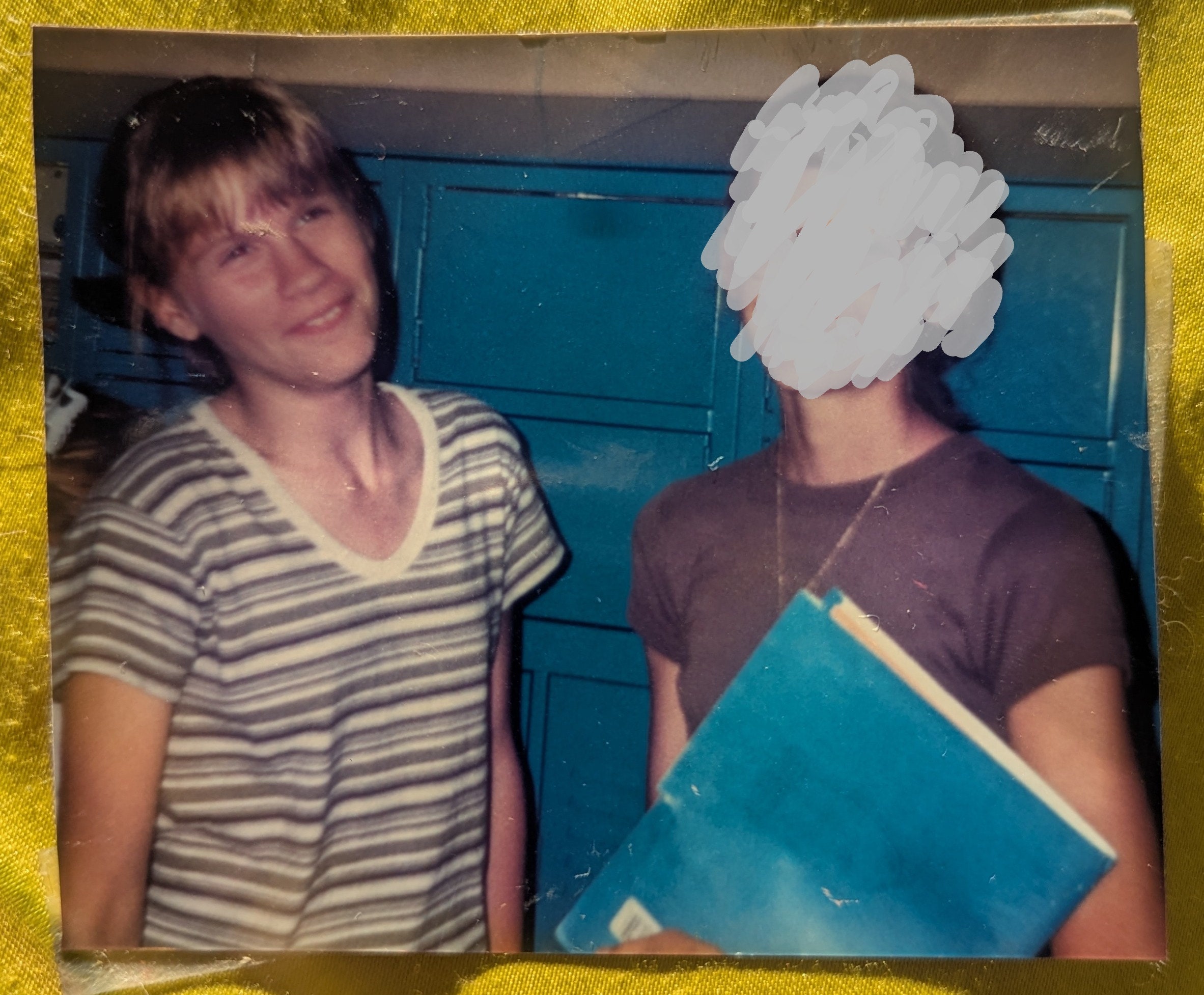 Photo of two middle school girls (one with her face blurred out). The one on the left has a dorky smile on her face, and is wearing a striped shirt with a v-neck. She has light brown bangs. The girls are standing in front of blue lockers.