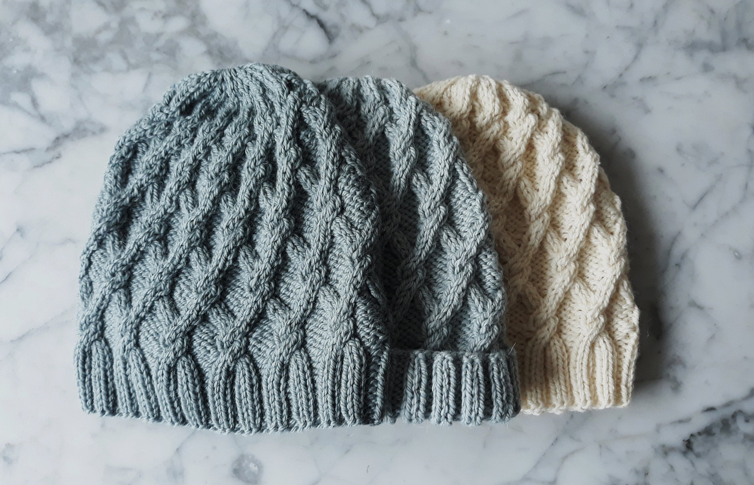 Three knitted cabled hats are laid flat, the hats are different colours: two are grey and one is creamy white.