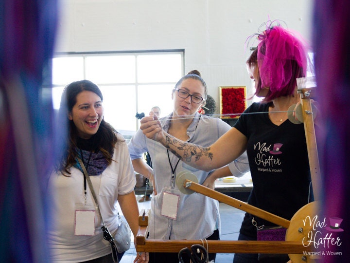 Queen Hatter is shown with two guests warping thread onto a wooden warping wheel. Queen Hatter has on a black shirt with the Mad Hatter logo on it, and a pink feathered hat. Queen Hatter and guests are looking at each other and smiling/ laughing.