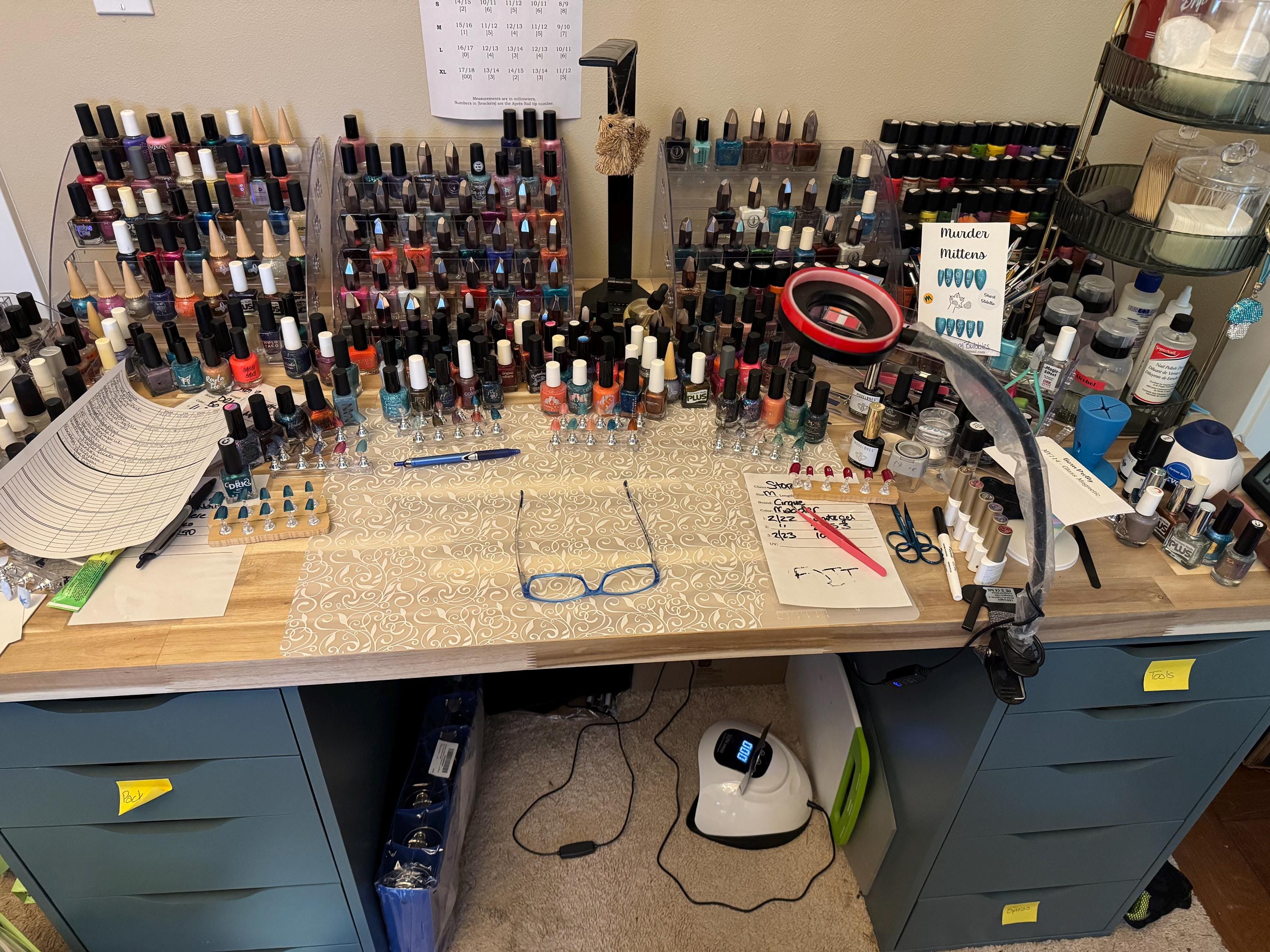 An extremely messy desk with over 4 racks of nail polish, polish bottles all over the desk, tools everywhere, with some almost finished projects