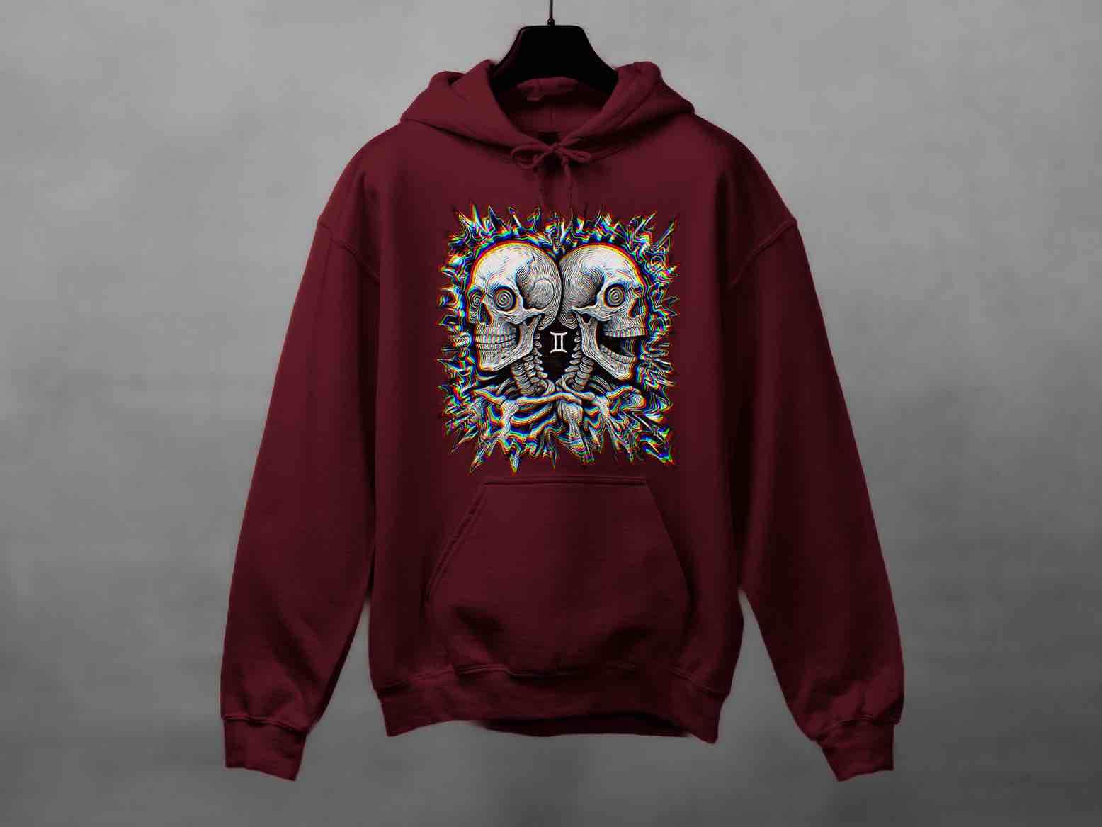Maroon hoodie displayed on a hanger against a gray wall. Large center print depicts back-to-back skulls with the Gemini “Ⅱ” and a neon glitch burst around them. Clear view of pocket, cuffs, and hood; zodiac skull graphic.