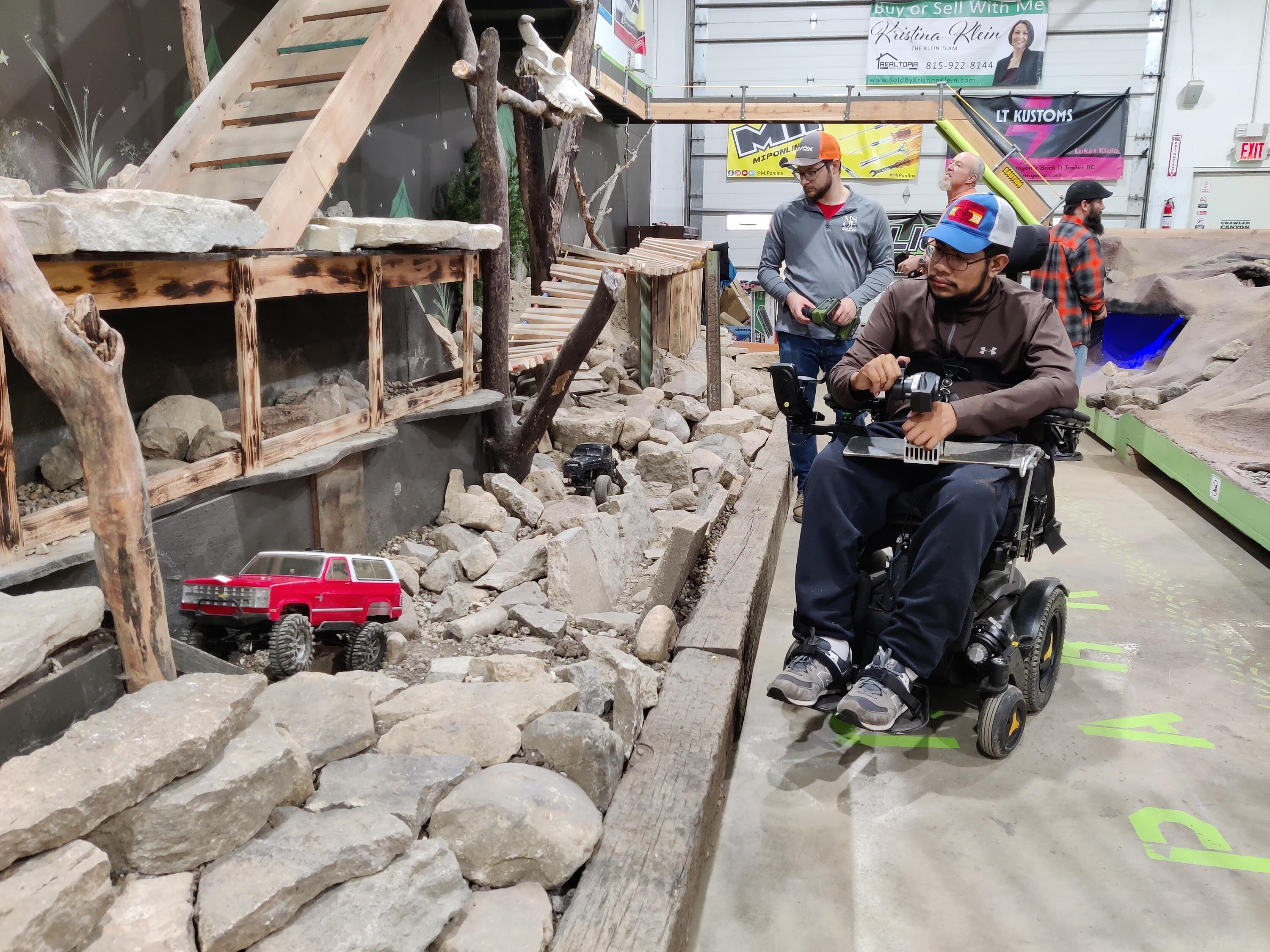 Individual in electric permobile wheelchair driving an RC car on a obstacle crawling course using adaptive equipment to control his transmitter