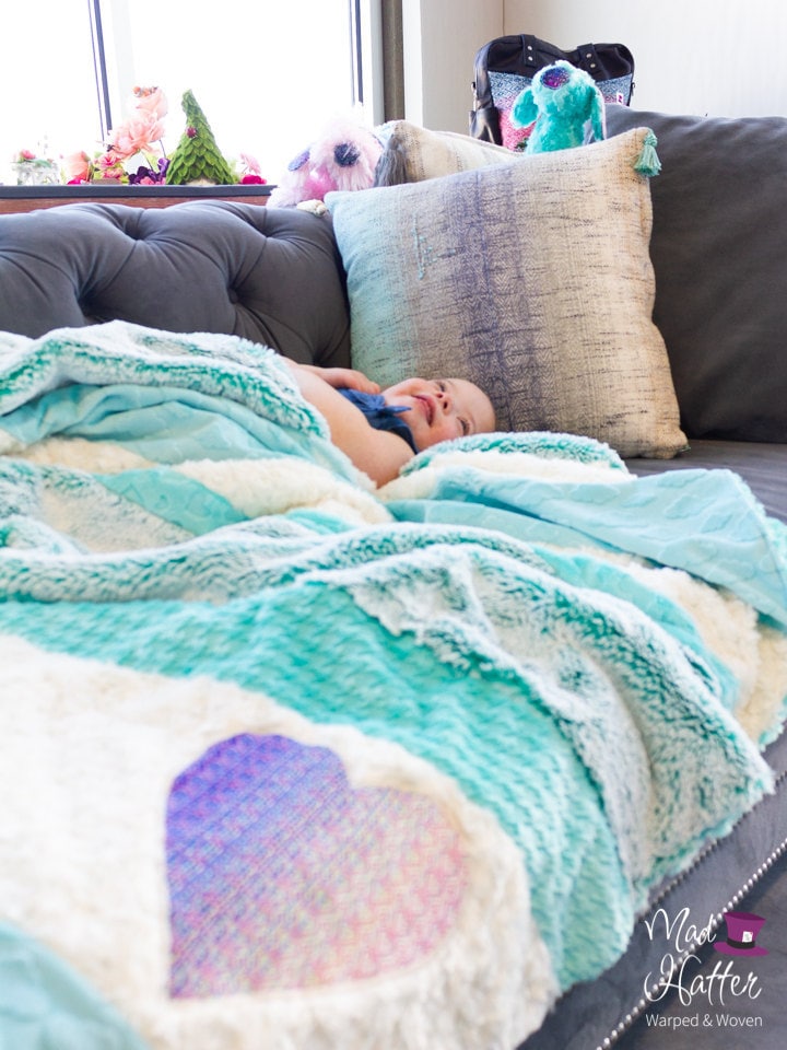 Young girl cuddled up in Mad Hatter Warped & Woven minky blanket on a grey couch.