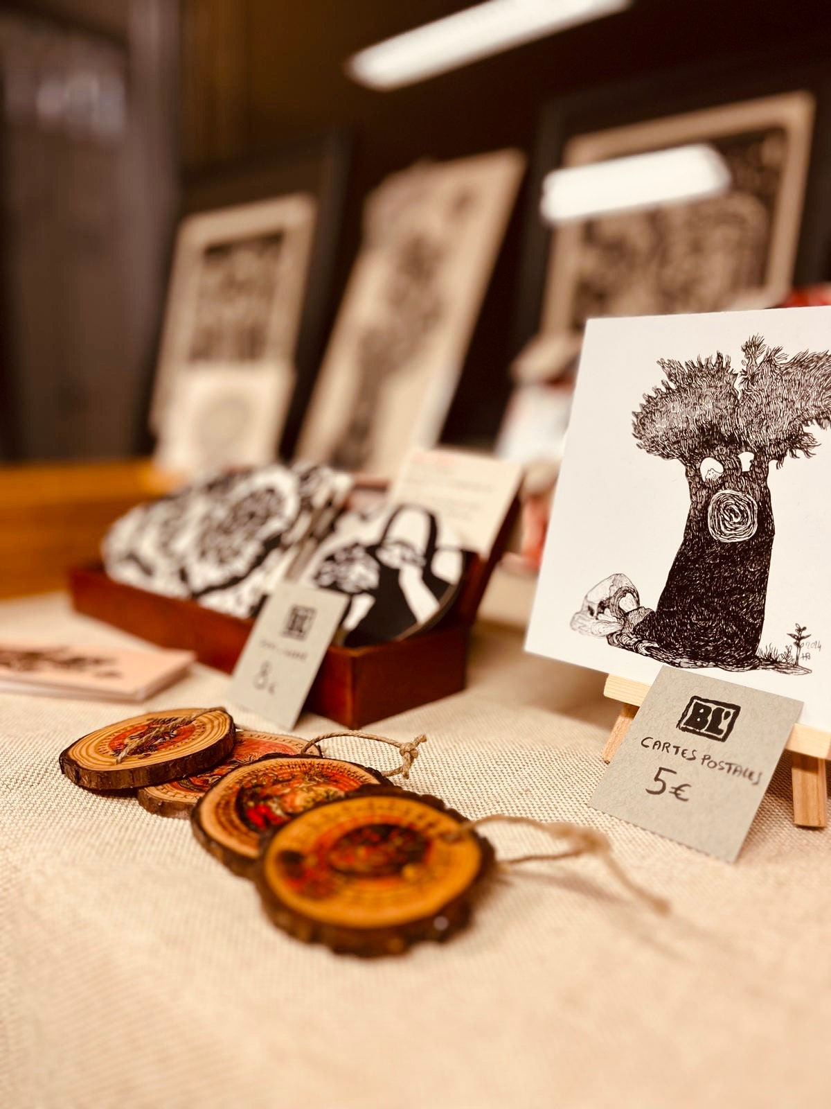 Détail des créations en linogravure, disposées sur un stand. Une carte postale représentant un arbre et des décorations en bois artisanales.