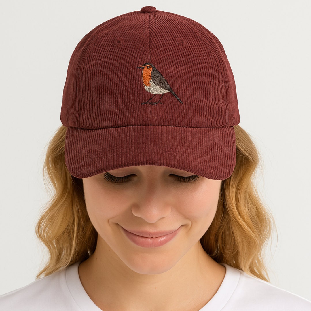 A close-up of a woman wearing a burgundy corduroy cap embroidered with a robin. She is smiling softly and looking down, photographed against a plain light background.