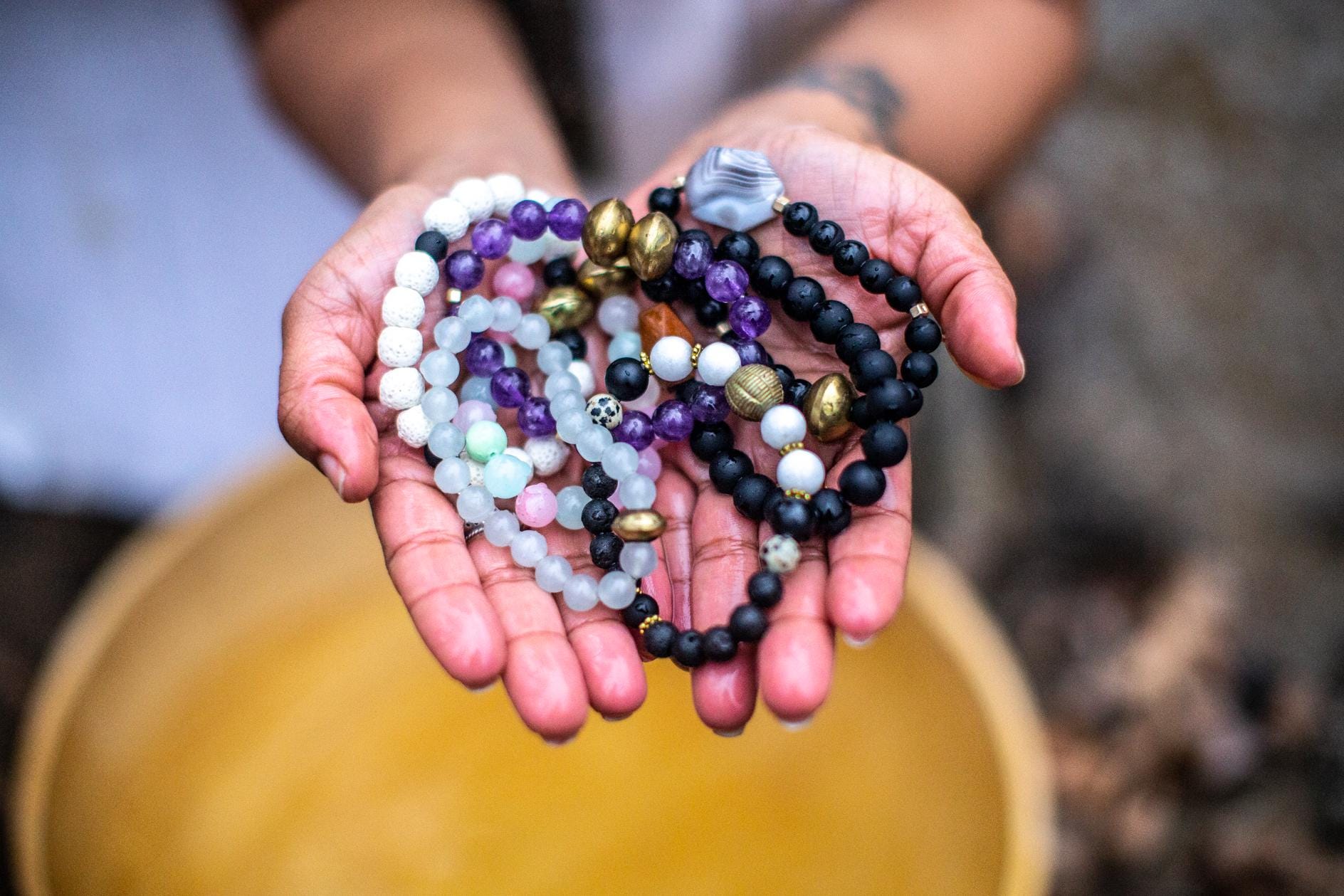 Hands holding assorted gemstone and beaded bracelets with amethyst, lava stone, and gold accents